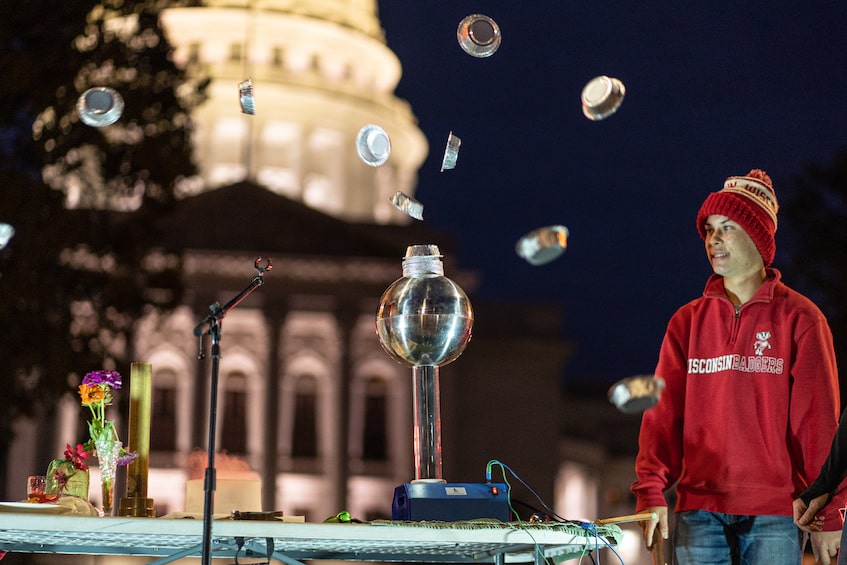 A man demonstrates a van de graf generator with pie pans. the WI capitol is in the background.