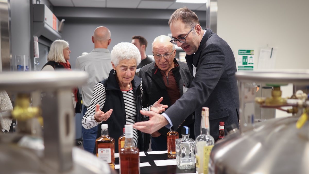 A research scientist and two invited guests look at an array of whiskey bottles set up on a lab bench for an experiment.