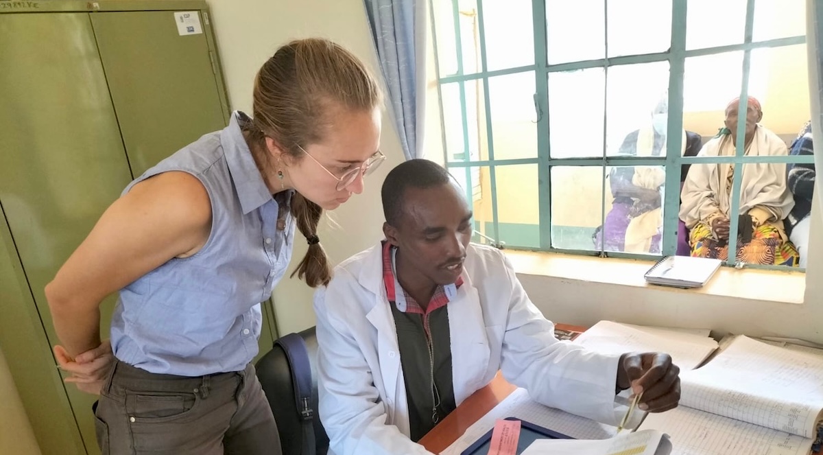 Kayla Huemer looks at papers over the shoulder of a man sitting at a desk in a clinical office in Africa.