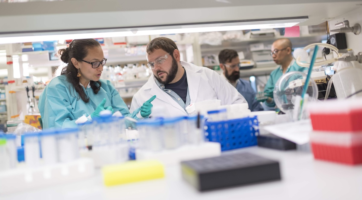 Scientists in white and blue lab coats sit in a lab space, a man and a woman sit in the foreground and two men stand in the background.