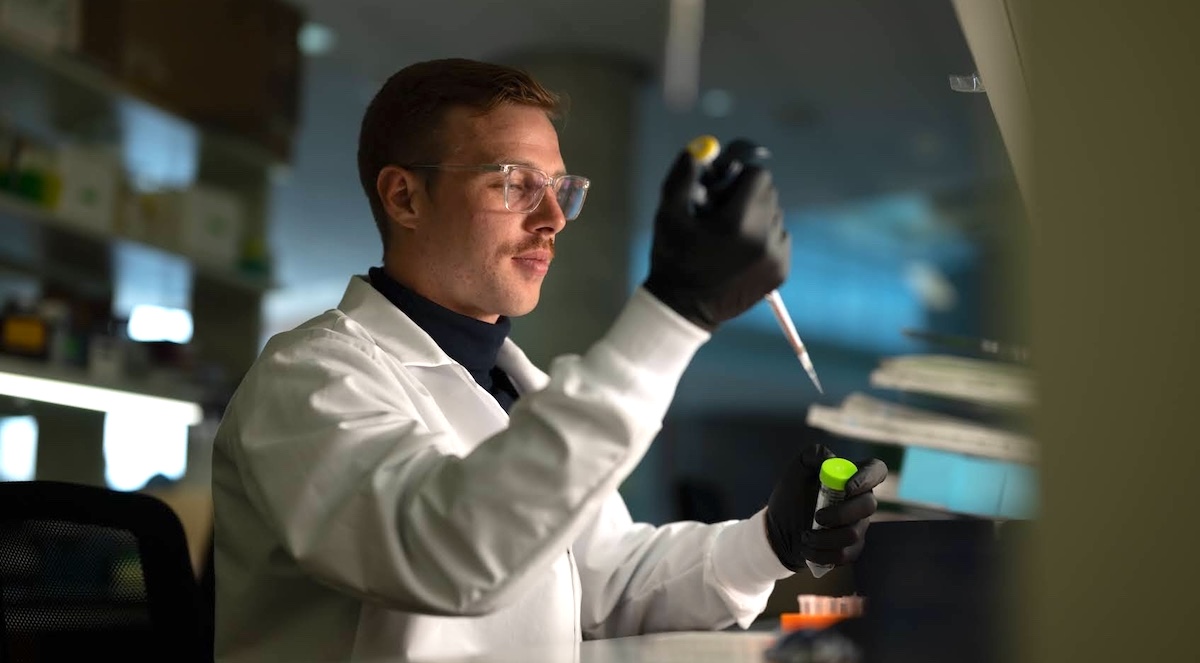A male scientist in a white lab coat and safety glasses sits in a darkened lab space. He is using a micropipette to transfer a sample into a tube.